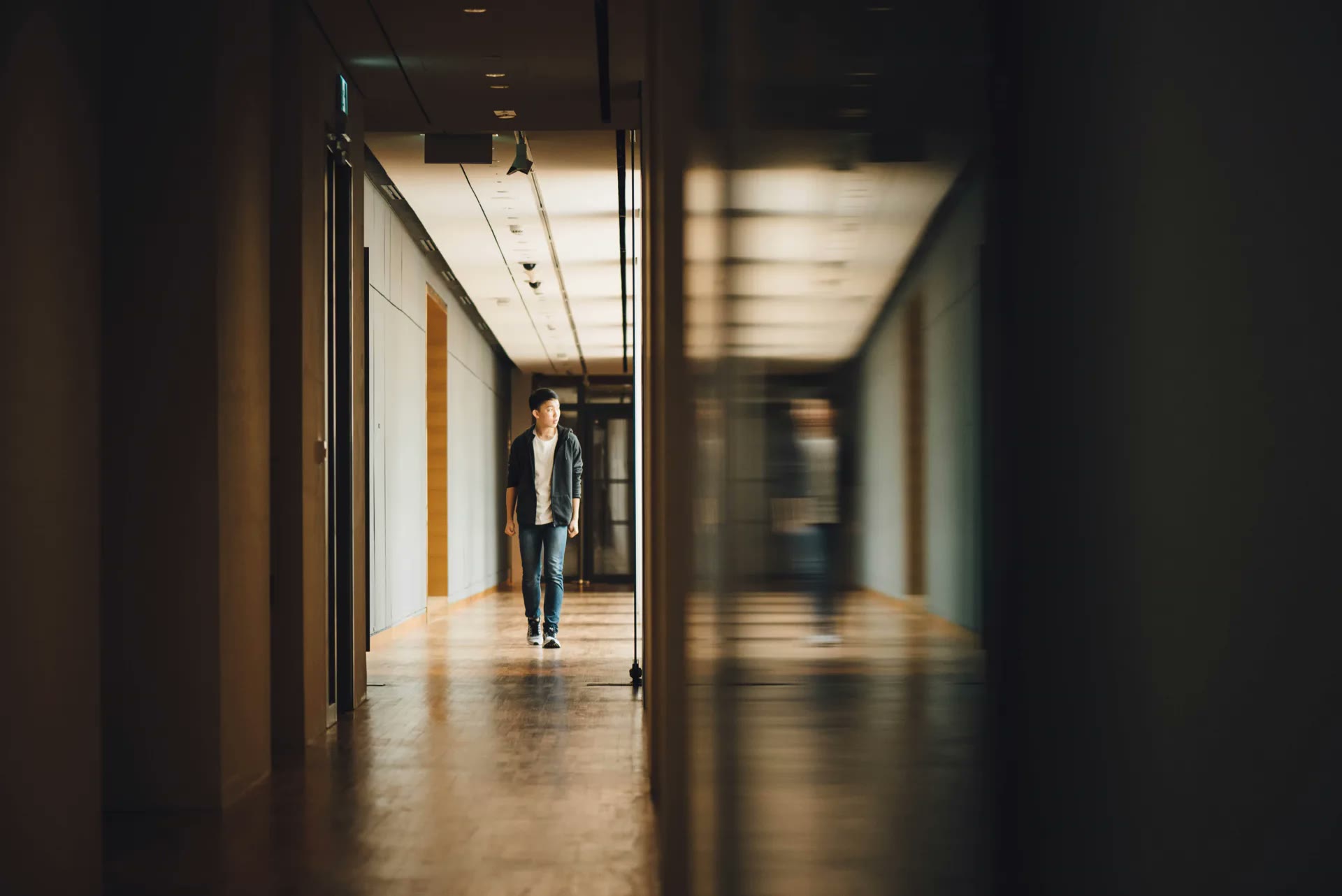 A person walking alone down a quiet office corridor toward a lit doorway, on the way to a meeting.