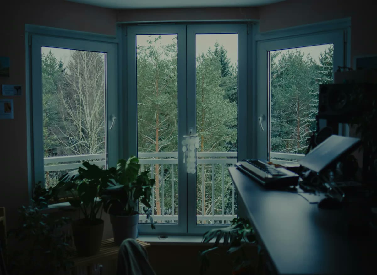 A quiet desk beside tall windows looking out on evergreen trees, a calm spot to pause and breathe.