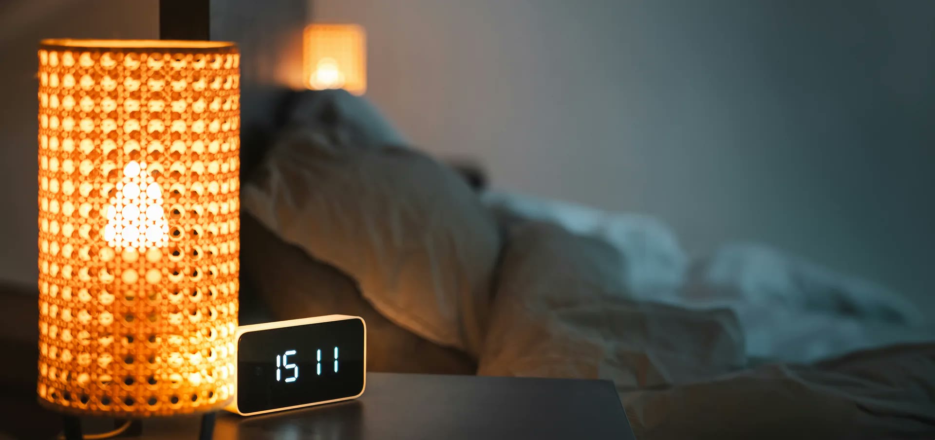 A digital clock glowing on a bedside table next to a warm lamp in a dark bedroom, late at night.