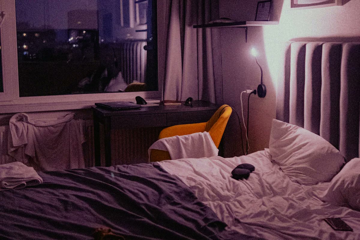 A dimly lit bedroom at night with an unmade bed, a soft reading light, and city lights visible through the window.