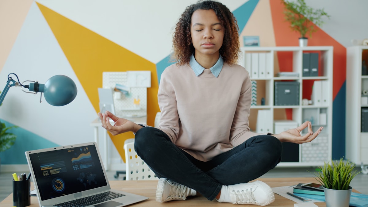 A woman pauses to meditate at her office desk beside an open laptop, illustrating a brief mindfulness break during a busy workday.