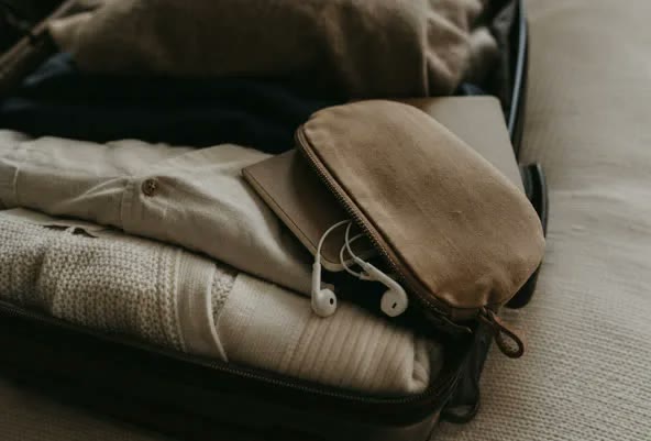 A small travel pouch with earbuds and a notebook tucked into the top of an open suitcase, ready to go