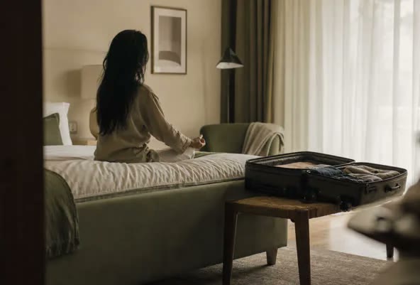 A person sitting cross-legged on a hotel bed, meditating, with an open suitcase nearby in soft morning light