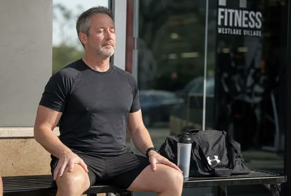 A man sitting on a bench outside a gym, gym bag at his feet, eyes closed and meditating peacefully after a workout